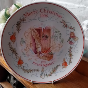 Puede incluir: Un plato de cerámica blanco con un diseño navideño de Peter Rabbit. El plato presenta una ilustración central de Peter Rabbit con una cesta de zanahorias, rodeado de imágenes festivas y el texto "Merry Christmas 1996 from Peter Rabbit."