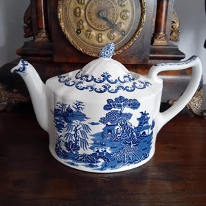 Puede incluir: Una tetera de cerámica blanca con un patrón azul de estilo willow que presenta ilustraciones detalladas de pagodas, árboles y un puente. La tetera incluye una tapa a juego con un pequeño remate, un pico curvo y un asa resistente con detalles.