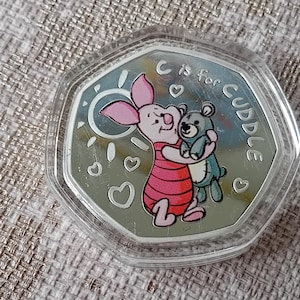 May include: A silver coin featuring Piglet hugging a teddy bear, with the text "C is for Cuddle." The coin is encased in a clear, hexagonal plastic case. The background is a textured, light brown surface.