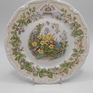 Puede incluir: Un plato de cerámica blanco con un diseño floral que presenta un ratón y un pájaro. El plato tiene un borde festoneado y dice "Brambley Hedge Spring" en letras marrones.
