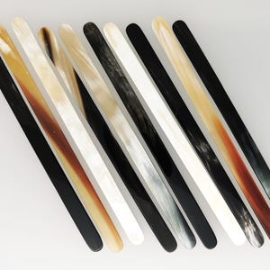 Puede incluir: Un surtido de diez palitos largos y delgados para remover, con extremos ovalados. Los palitos varían en color, incluyendo negro, blanco, beige y tonos de marrón. Están dispuestos diagonalmente sobre un fondo blanco, mostrando sus diversos tonos y texturas.