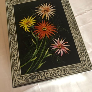 May include: A vintage silver-colored box with an ornate floral design. The box features a black background with a bouquet of five flowers in shades of pink, yellow, orange, and red.