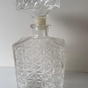 Puede incluir: Una jarra de cristal transparente con un patrón geométrico y un tapón a juego. El tapón es rectangular con un diseño facetado. La jarra tiene una base cuadrada y un tapón de corcho.