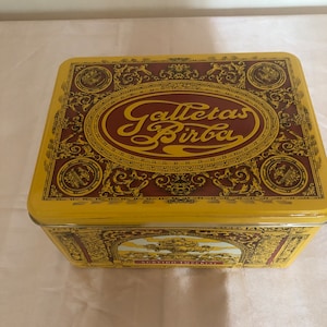 Puede incluir: Una lata rectangular amarilla con detalles ornamentados en marrón y dorado. La tapa presenta el texto "Galletas Birba" en una fuente dorada estilizada dentro de un marco ovalado. La lata es probablemente para galletas.