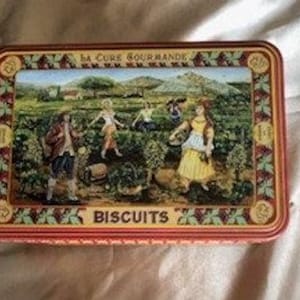 Op de afbeelding: Een vintage blikken doos met een kleurrijke illustratie van mensen die druiven oogsten in een wijngaard. De tekst "La Cure Gourmande" en "Biscuits" staat op de doos gedrukt.
