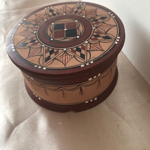 Peut inclure: Une boîte ronde en bois brun avec un motif peint. Le dessus de la boîte présente un motif géométrique dans des tons de brun, noir et blanc. Le bas de la boîte a une bordure blanche et brune avec un motif de frange.