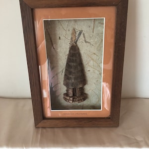 以下が含まれることがあります： ニューカレドニアの伝統的な部族の工芸品を模した木製の彫刻が額装されています。彫刻は自然素材で作られており、質感のある背景に飾られています。フレームは茶色の木で、前面はガラス製です。フレームの下部には「ヌーベル・カレドニ」という文字が印刷されています。