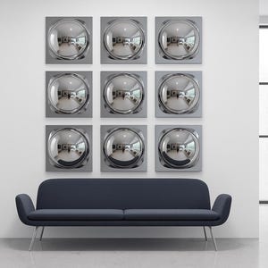 Op de afbeelding: Negen vierkante wandkunstwerken met een reflecterend, zilverkleurig, cirkelvormig ontwerp zijn gemonteerd boven een donkerblauwe bank met zilveren poten. De kamer heeft grote ramen en een minimalistische esthetiek.
