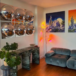 May include: A room interior featuring a wall-mounted grid of six circular convex silver mirrors, a green marble console table with arched legs, a blue tufted sofa, a tall floor lamp with multi-coloured light projections, and desert landscape art.