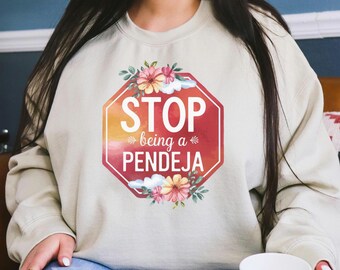 Może przedstawiać: Beżowa bluza z czerwoną grafiką znaku stopu. Znak zawiera białe napisy "STOP" i "being a PENDEJA", z kwiatowymi i chmurowymi akcentami. Bluza ma swobodny styl.