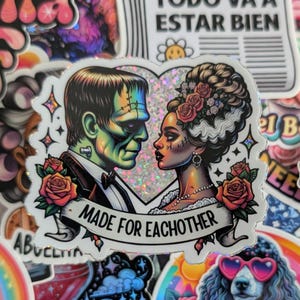 May include: A sticker featuring a couple, with the Frankenstein monster and his bride, in a heart-shaped design. The sticker has a banner that reads "MADE FOR EACHOTHER" with roses on each side. The background is a glittery silver.