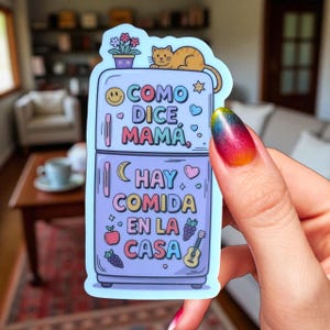 May include: A sticker in the shape of a refrigerator with the text "COMO DICE MAMA" and "HAY COMIDA EN LA CASA". The sticker has a cat on top, flowers, and various food-related illustrations.