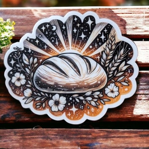 May include: A white sticker with a black and white illustration of a loaf of bread surrounded by flowers and wheat stalks. The bread is illuminated by a golden light with stars and other celestial objects in the background.