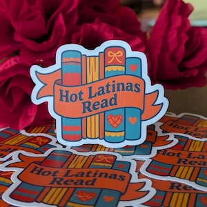 May include: A sticker featuring a stack of books with a banner that reads "Hot Latinas Read." The books are blue, orange, and yellow with heart and bow details. The sticker is on a surface with other stickers and red flowers.