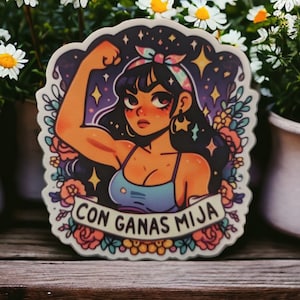 May include: A sticker featuring a cartoon woman with dark hair, wearing a blue tank top and a pink and blue bandana. She is flexing her bicep and has a determined expression. The background is a dark blue with stars and the text "CON GANAS MIJA" is on a banner.