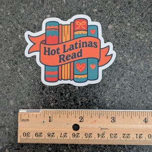 May include: A sticker featuring a stack of books with the text "Hot Latinas Read" on an orange banner. The books are teal, orange, and red with heart and bow details. A ruler is at the bottom, measuring in inches.