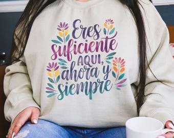 May include: A light beige sweatshirt with the text "Eres suficiente aqui, ahora y siempre" in purple, blue, and yellow lettering with floral accents.