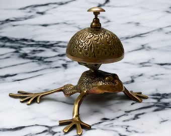 May include: A brass frog figurine with a bell on its back. The bell has a decorative design and a small knob on top. The frog is sitting on a white marble surface.