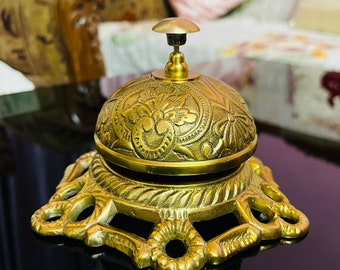May include: A gold-colored brass bell with an ornate design. The bell has a round top and a base with a decorative pattern. The bell is sitting on a black surface.