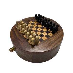 Op de afbeelding: Een houten schaakset met een ronde, scharnierende doos. Het bord is open en toont zwarte en witte vakjes met zwarte en witte schaakstukken.