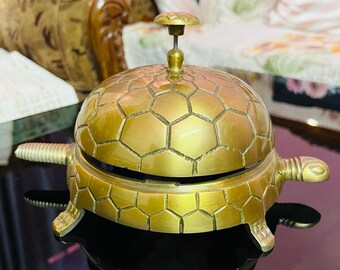 May include: A gold-coloured turtle-shaped bell with a hexagonal pattern on the top and bottom. The bell is sitting on a black surface.