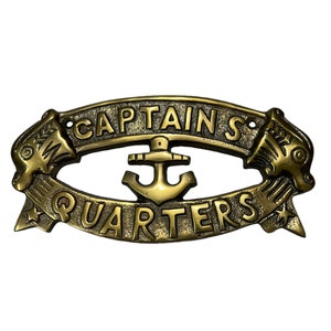 Pode incluir: Um sinal de metal dourado com tema náutico. O sinal diz "CAPTAINS QUARTERS" e apresenta uma âncora no centro. O sinal é decorado com dois peixes estilizados de cada lado.