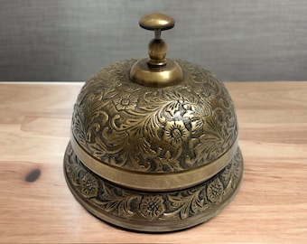 May include: A gold-toned brass service bell with an ornate floral design. The bell has a rounded top and a base with a decorative pattern.