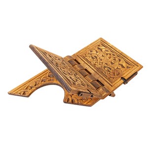 May include: A wooden book stand with intricate carved designs. The stand is made of light brown wood and features detailed patterns on the front and sides. The stand is open, with the pages angled for easy reading. The hinges are visible, and the overall design is ornate.