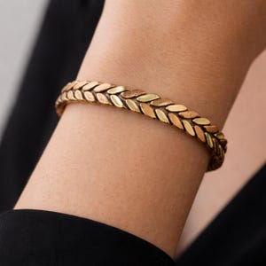 May include: A gold-toned bracelet with a woven design. The bracelet features a series of leaf-shaped links in alternating shades of gold and copper. The jewelry is worn on a wrist, with a black garment in the background.