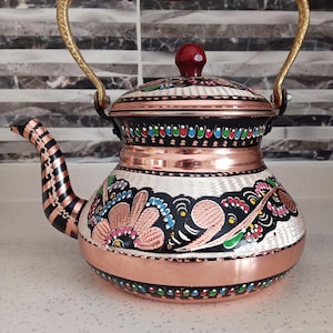 May include: A traditional Turkish copper teapot with a floral design. The teapot has a black and white floral pattern with pink, blue, green, and yellow accents. The teapot has a copper handle and a copper lid with a wooden knob.