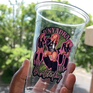 以下が含まれることがあります： 透明なプラスチック製の使い捨てカップで、カスタムグラフィックデカールが施されています。黒い衣装を着た人物の画像3枚、緑のヤシの葉のアクセント、そして「D'NYADRA」と「YOU DA Birthday」というピンクと黒の文字がデザインされています。