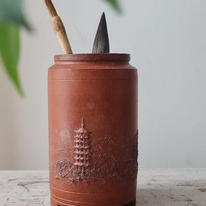 Peut inclure: Un porte-pinceaux cylindrique en céramique brun rougeâtre avec un relief détaillé d'une pagode et d'un paysage. Deux pinceaux de calligraphie sont insérés dans le porte-pinceaux. Le porte-pinceaux est sur une surface en bois blanc vieilli.