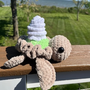 May include: A crocheted turtle toy with a white and green shell. The turtle is sitting on a wooden railing with a body of water and green grass in the background.