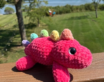 May include: A pink crocheted dinosaur toy with rainbow colored pom-poms on its back. The toy has black eyes and a friendly expression.