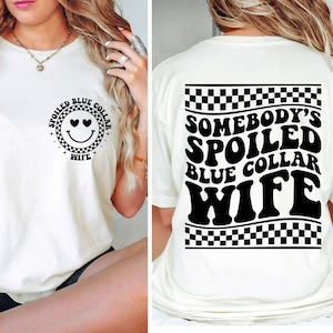 May include: White t-shirt with a black and white checkered design and the text "Somebody's Spoiled Blue Collar Wife" on the back. The front of the shirt has a black and white smiley face with hearts for eyes and the text "Spoiled Blue Collar Wife" in a circle.
