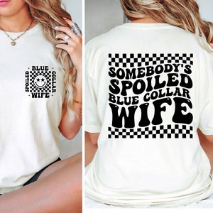 Könnte beinhalten: Weißes T-Shirt mit einem schwarz-weißen Karomuster auf der Rückseite. Der Text auf der Rückseite lautet "Somebody's Spoiled Blue Collar Wife". Die Vorderseite des Shirts hat ein schwarz-weißes Karomuster mit dem Text "Spoiled Blue Collar Wife" und einem Smiley.