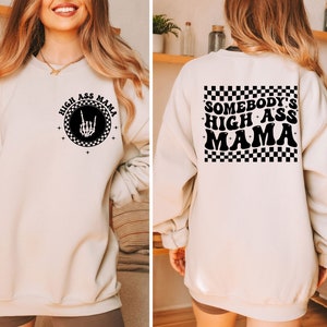 Puede incluir: Una sudadera de color crema con un gráfico negro en la parte delantera que dice "High Ass Mama" con una mano de esqueleto haciendo un signo de rock and roll. La parte posterior de la sudadera tiene un patrón de cuadros blanco y negro con el texto "Somebody's High Ass Mama" en negro.