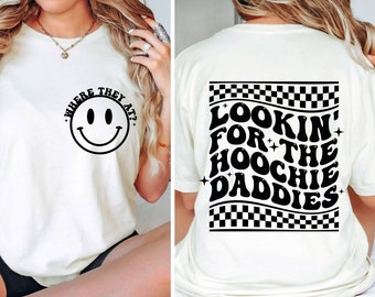 Pode incluir: Camiseta branca com um rosto sorridente preto e o texto "Where they at?" na frente e o texto "Lookin' for the hoochie daddies" com um padrão xadrez nas costas.