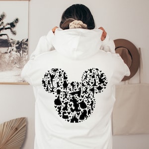 Puede incluir: Sudadera blanca con una silueta negra de la cabeza de Mickey Mouse formada por varios personajes de Disney.