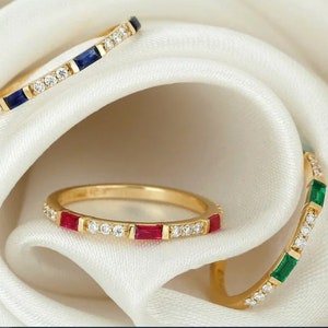 Könnte beinhalten: Drei goldene Ringe mit einer Reihe kleiner Diamanten und einem größeren farbigem Edelstein in der Mitte. Die Ringe sind blau, rot und grün.