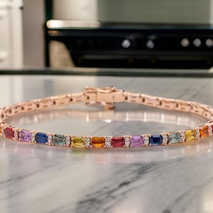 May include: A rose gold bracelet with a row of colorful gemstones. The gemstones are set in a rectangular shape and are surrounded by smaller diamonds. The bracelet is on a white marble surface.