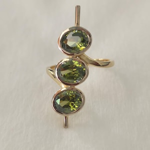 以下が含まれることがあります： 3つの楕円形オリーブグリーンの宝石をあしらったゴールドリング。宝石は垂直で流れるようなデザインにセットされています。リングは研磨仕上げで、ユニークでモダンなスタイルです。ジュエリーです。