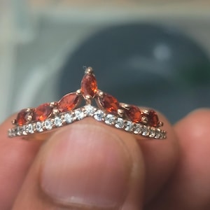 Op de afbeelding: Een roségouden ring met een chevron-ontwerp. De ring heeft een rij kleine, heldere stenen aan de onderkant en een rij oranjerode marquise-geslepen stenen aan de bovenkant, waardoor een V-vorm ontstaat. De ring wordt door een persoon vastgehouden.