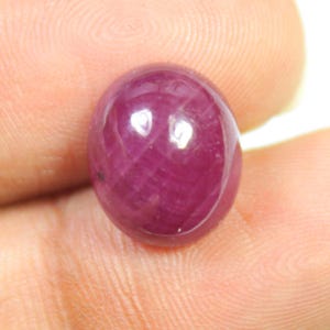 Könnte beinhalten: Ein polierter, ovalförmiger Rubin-Edelstein. Der Edelstein hat eine tiefe, satte rote Farbe mit einer glatten, reflektierenden Oberfläche. Der Rubin hat eine leichte Transluzenz. Der Edelstein wird in einer Hand gehalten.