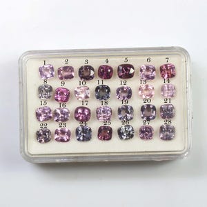 Könnte beinhalten: Eine Sammlung von 28 quadratisch geschliffenen Edelsteinen in einer transparenten Kunststoffbox. Die Edelsteine variieren in den Farbtönen Lila, Rosa und Grau. Jeder Stein ist von 1 bis 28 nummeriert und in Reihen und Spalten angeordnet.