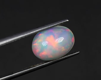 Puede incluir: Una piedra preciosa de ópalo ovalada blanca con colores arcoíris iridiscentes, sostenida por pinzas sobre un fondo negro.