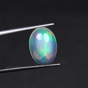 Può includere: Una pietra preziosa di opale ovale bianca con bagliori verdi e arancioni. L'opale è tenuta da una pinzetta.