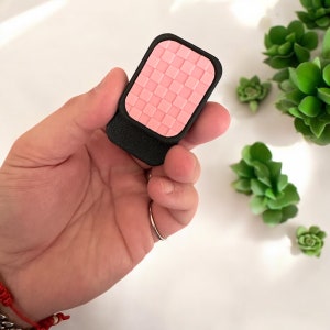 May include: A small, pink and black square-shaped object with a textured surface. The object is held in a hand.