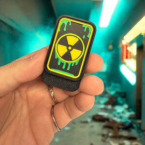 May include: A black patch with a yellow and green radioactive symbol and dripping green paint. The patch is attached to a black velcro backing.