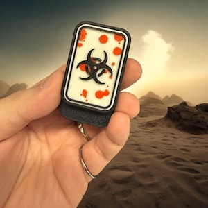 May include: A black and white biohazard symbol with red splatter on a white background. The symbol is on a black plastic patch.